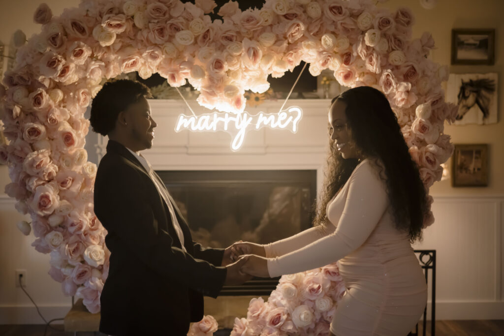 Chris and Kiki hold hands in front of a pink floral heart shaped arch and a sign that says "marry me"