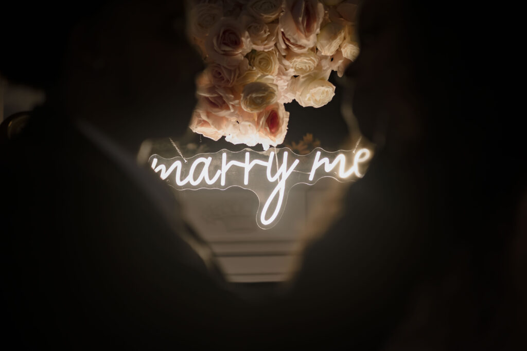 Chris and Kiki are blurred out in the foreground while the camera focuses on the "marry me" sign.